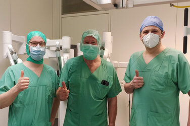 Dr. med. Yannick Lippka, Dr. med. Georg Schön und Dr. med. Marcel Stoll stehen in OP-Kleidung und recken den Daumen empor. 