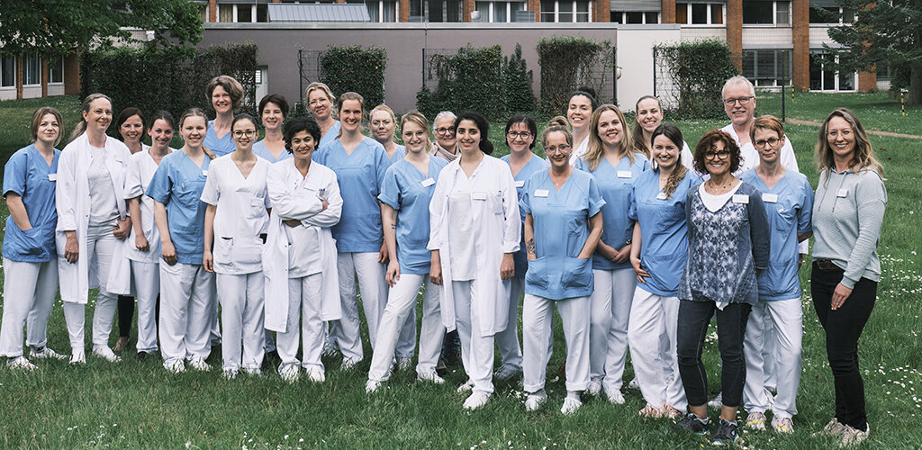 Die Mitarbeiter*Innen des Brustzentrum KRH Klinikum Robert Koch Gehrden stehen lächelnd auf einer grünen Wiese vor dem Klinikkomplex. 